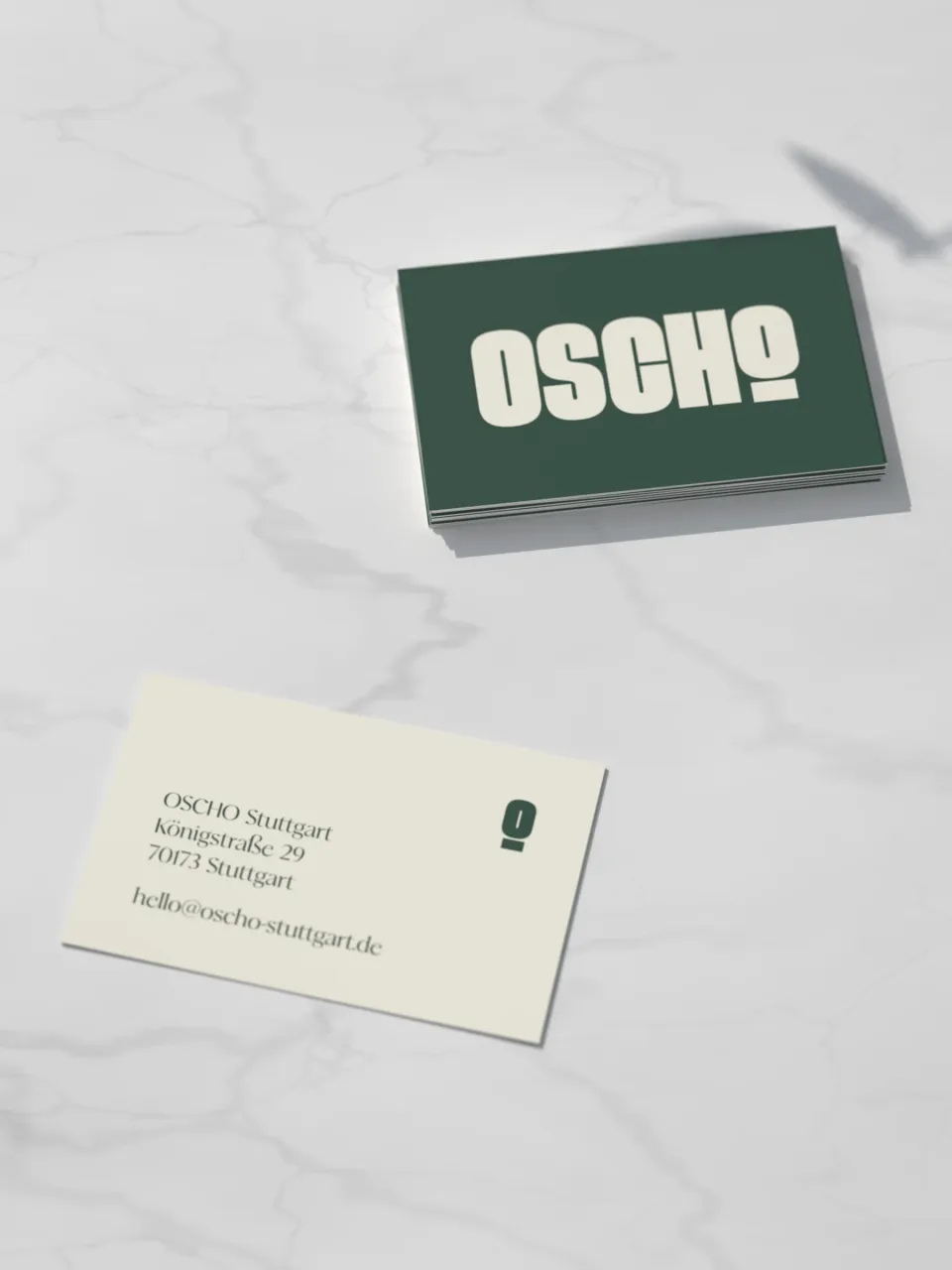 OSCHO: Visitenkarten auf Marmor – vorn die Wortmarke in Creme auf Dunkelgrün, hinten Adresse und Bildmarke auf Creme